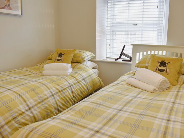 Ein Zimmer mit zwei Betten, jeweils mit gelb-kariertem Bettbezug und Kissen mit Bienenmuster. Es gibt ein Fenster mit Jalousien, einen Nachttisch mit Lampe und ein gerahmtes Bild an der Wand.