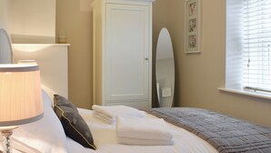 Een net opgemaakt bed met witte lakens en een grijze deken, een bedlampje, een spiegel en een raam met zonwering.