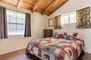 Une chambre avec un plafond en bois, un grand lit recouvert d’une courtepointe à motifs, deux fenêtres avec des rideaux et un cadre photo fixé au mur.