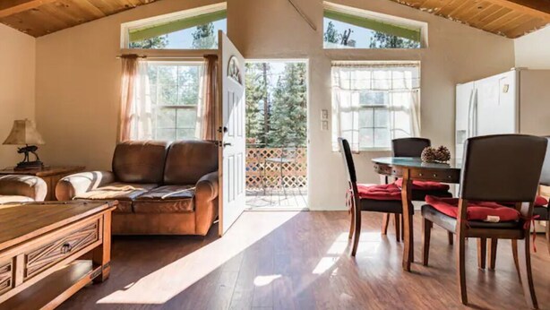 A cozy living room with a wooden ceiling, a leather sofa, a dining table with chairs, and a view of the outdoors through large windows. - Cute Cozy Cabin- View of the Slopes-Pets OK! (Big Bear Lake)