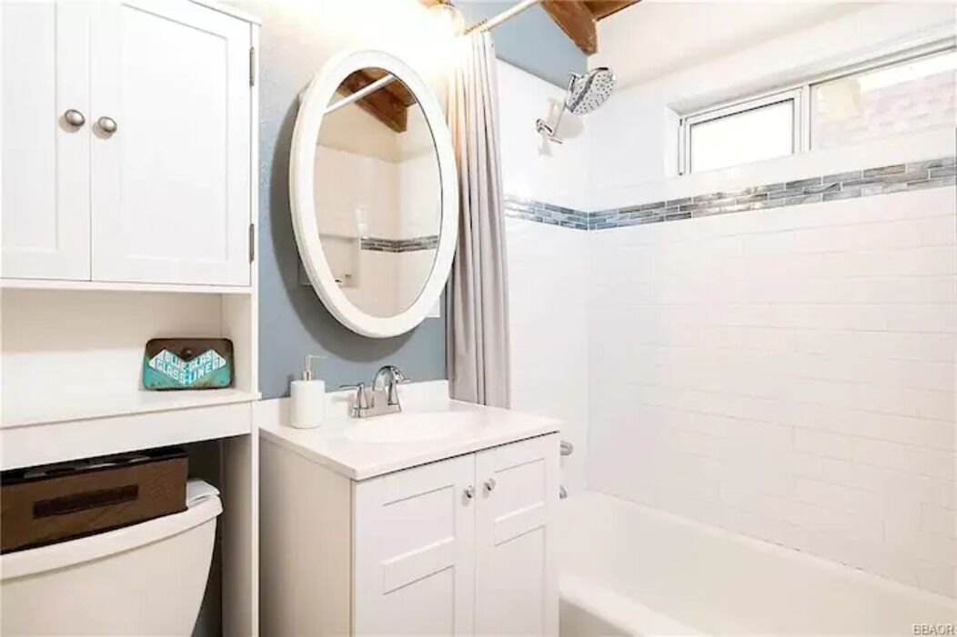 A bathroom with a white sink, mirror, and cabinet, a shower, and a window above the shower.