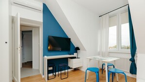 Una habitación moderna con techo inclinado, un escritorio con televisor de pantalla plana, dos sillas azules y una ventana con cortinas.