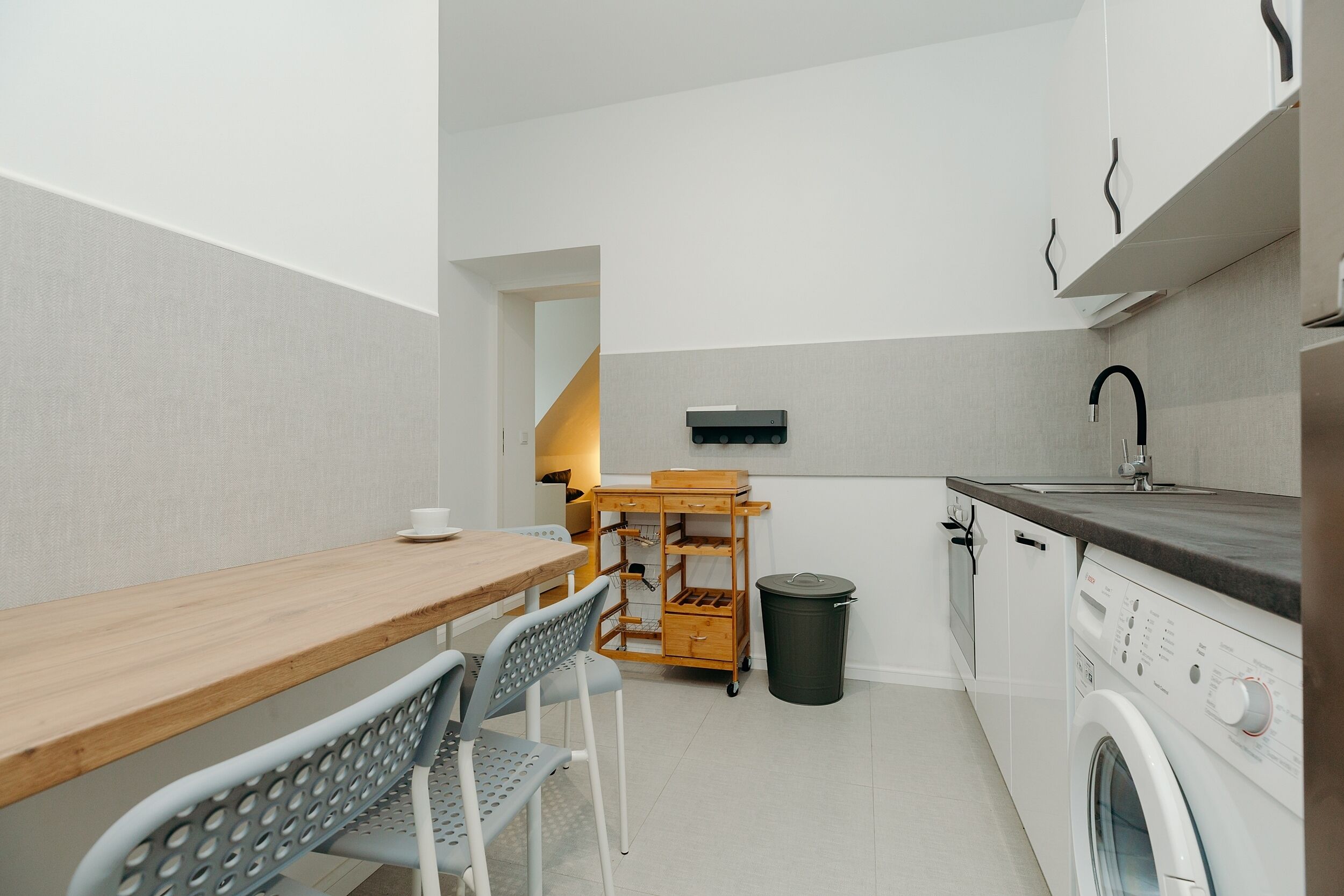 Une cuisine compacte avec une table en bois, deux chaises, un petit chariot en bois, un lave-linge et une poubelle.