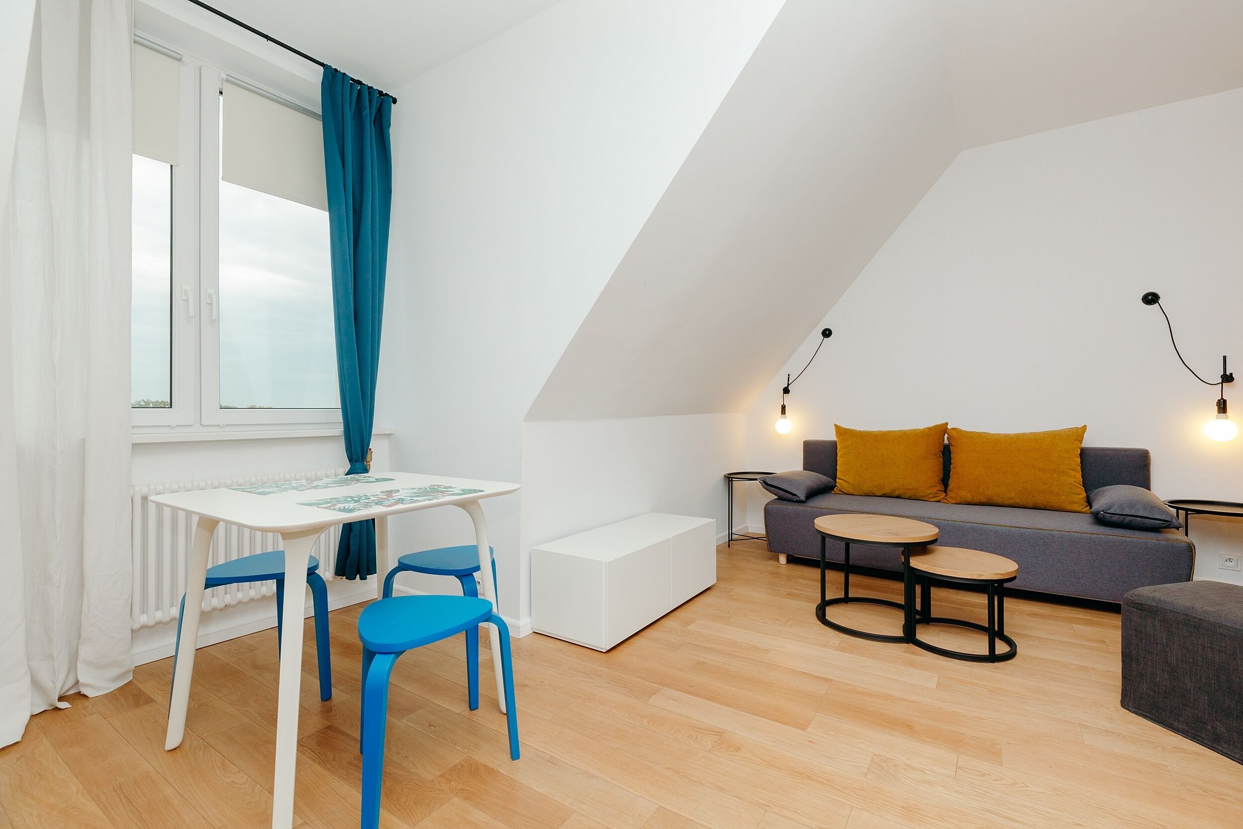 A modern, well-lit room with a white table, blue chairs, a grey sofa with yellow cushions, and a small round table.