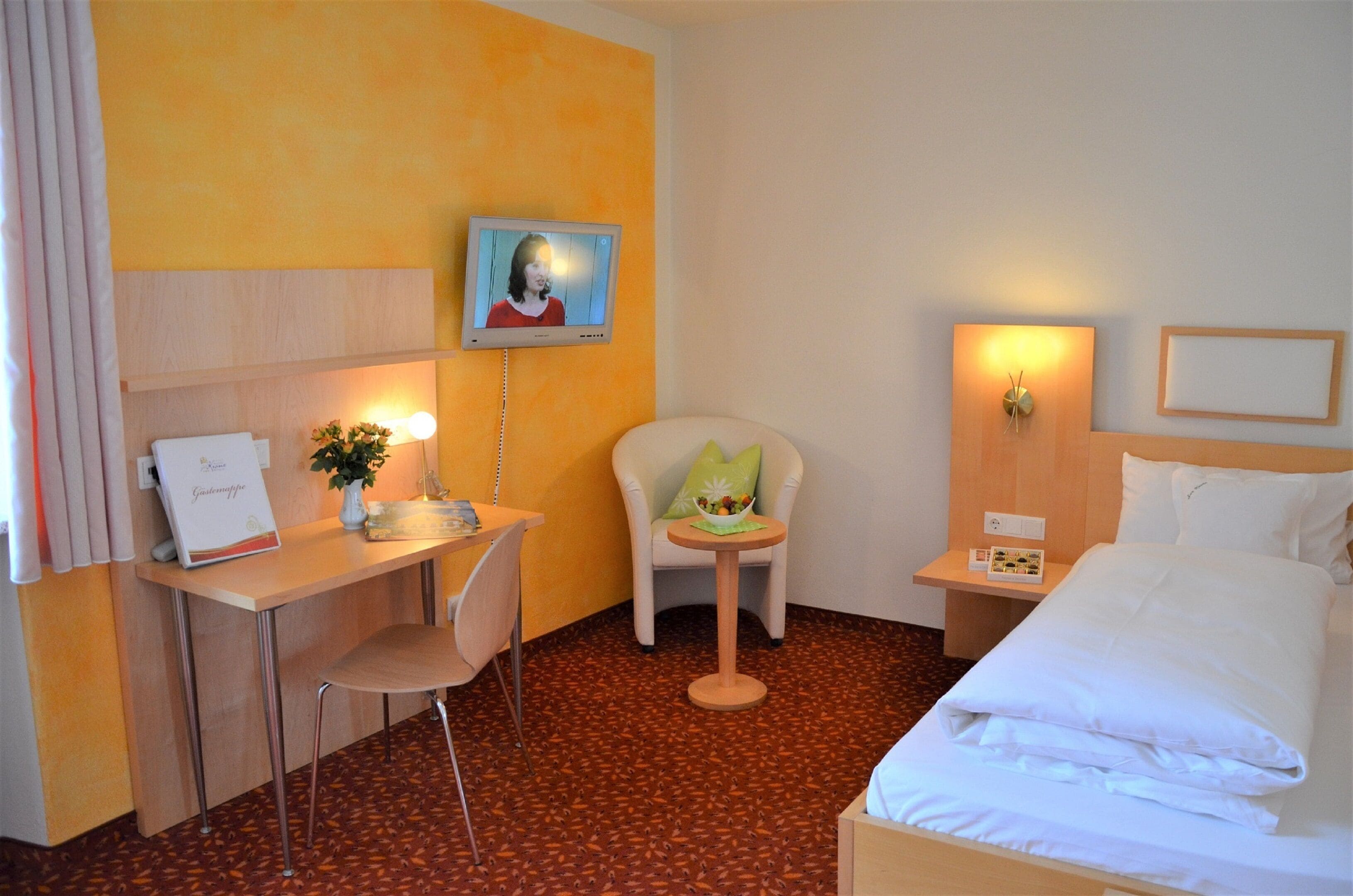 A hotel room with a bed, a desk with a chair, a small table with a chair, a wall-mounted TV, and a bedside table with a lamp.