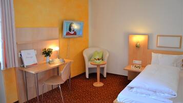 A hotel room with a bed, a desk with a chair, a small table with a chair, a wall-mounted TV, and a bedside table with a lamp.