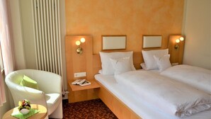 Hotellihuone, jossa on sänky, kaksi yöpöytää, tuoli, pieni pöytä, jonka päällä on kulho täynnä hedelmiä, ja patteri.