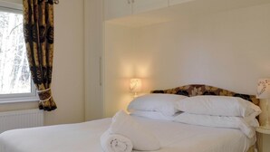 A neatly made bed with white linens and a rolled towel, a bedside lamp, and a window with curtains.