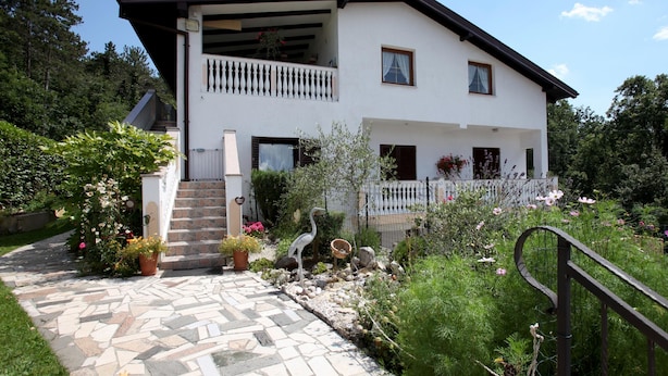 Ein zweistöckiges Haus mit einem gepflasterten Eingangsbereich, einem Balkon mit weißer Geländer und einem Garten mit verschiedenen Pflanzen und Blumen.