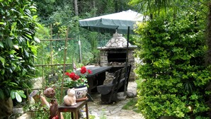 Eine Steinküche im Freien mit Pizzaofen, umgeben von üppiger Vegetation und einem kleinen Tisch mit einer Katzenfigur.