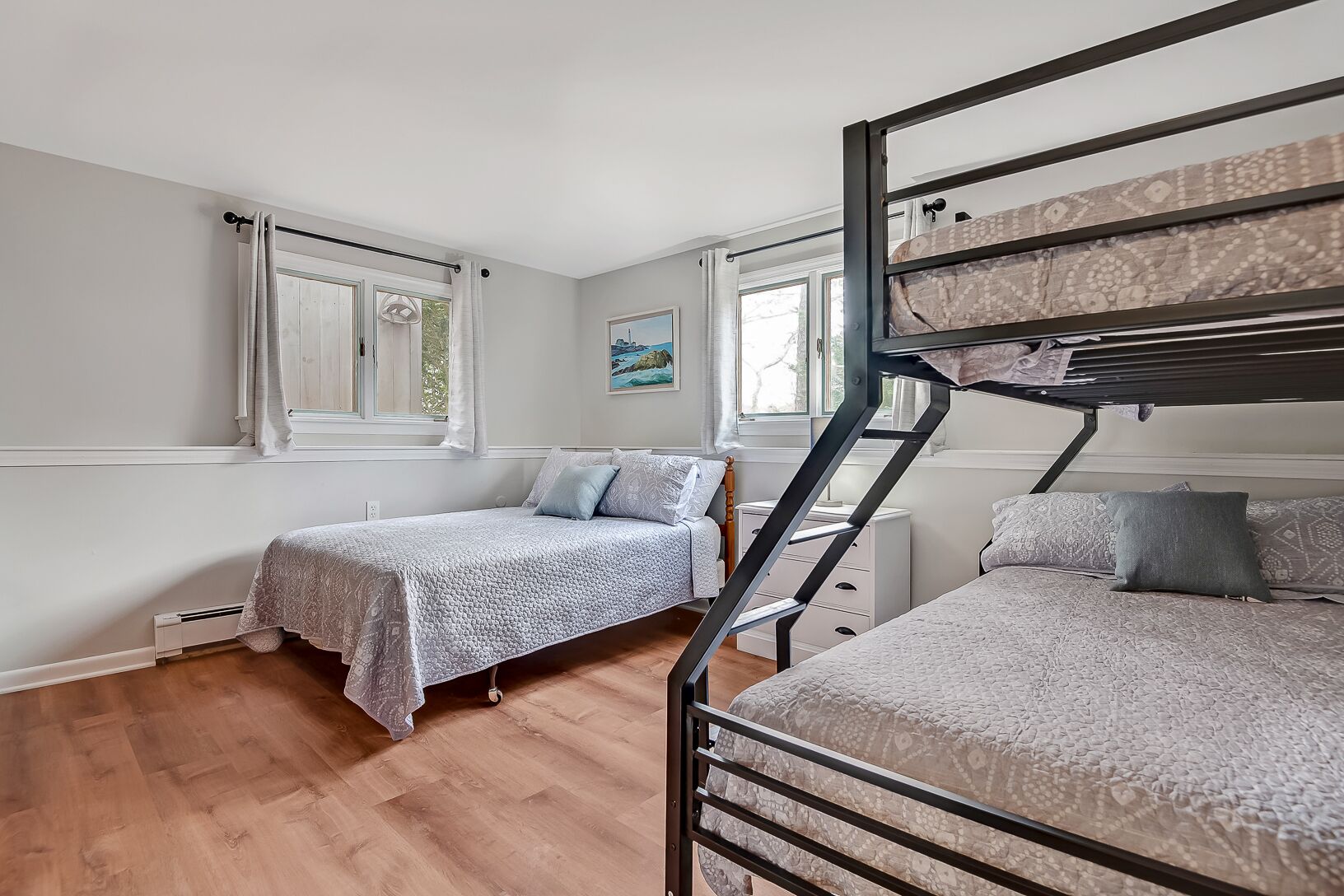A dormitory room with two single beds, a bunk bed, a small white dresser, and a window with curtains.