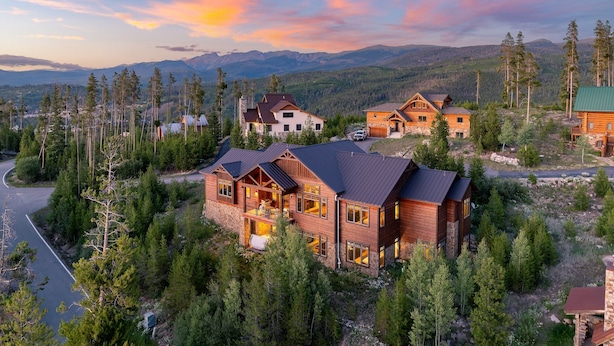 Une grande cabane en rondins dotée de plusieurs chambres et d’un balcon, entourée d’une forêt dense et de montagnes.