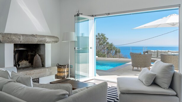 Un soggiorno moderno con camino, un’area salotto a bordo piscina e una porta vetrata che si apre su un balcone con vista sul mare.