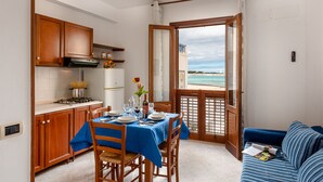 A compact kitchen with wooden cabinets, a dining table set for two, and a view of the beach through an open door. - I Tre Golfi Beach Apartments (San Vito Lo Capo)