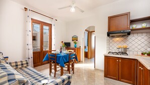 A cozy living room with a blue sofa and a dining area with a table set for two. A kitchen with a stove and cabinets is visible in the background. - I Tre Golfi Beach Apartments (San Vito Lo Capo)
