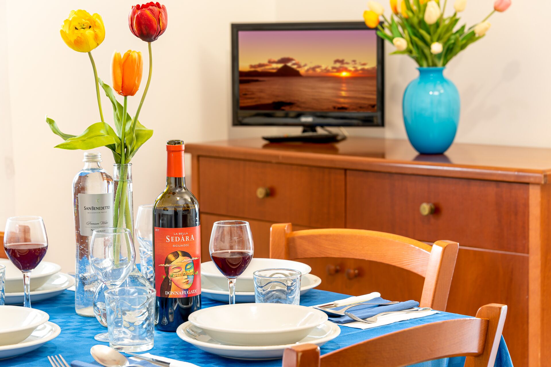 A dining table set with plates, glasses, and a bottle of wine. There are tulips in a vase and a television displaying a sunset.