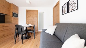 A modern, compact living space with a grey sofa, wooden cabinets, a dining table with chairs, and a radiator. - Al Forte 6 - One Bedroom Apartment, Sleeps 3 (Valdisotto)