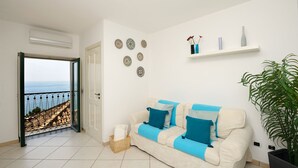 A living room with a beige sofa, blue cushions, a potted plant, decorative plates, and a view of the sea through a balcony door. - Ananas - Sea View - Fruit Flats by AMALFIVACATION.IT (MINORI)