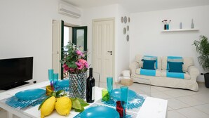 A living room with a TV, a dining table set with blue plates and glasses, a vase with flowers, and a sofa with blue cushions. - Ananas - Sea View - Fruit Flats by AMALFIVACATION.IT (MINORI)
