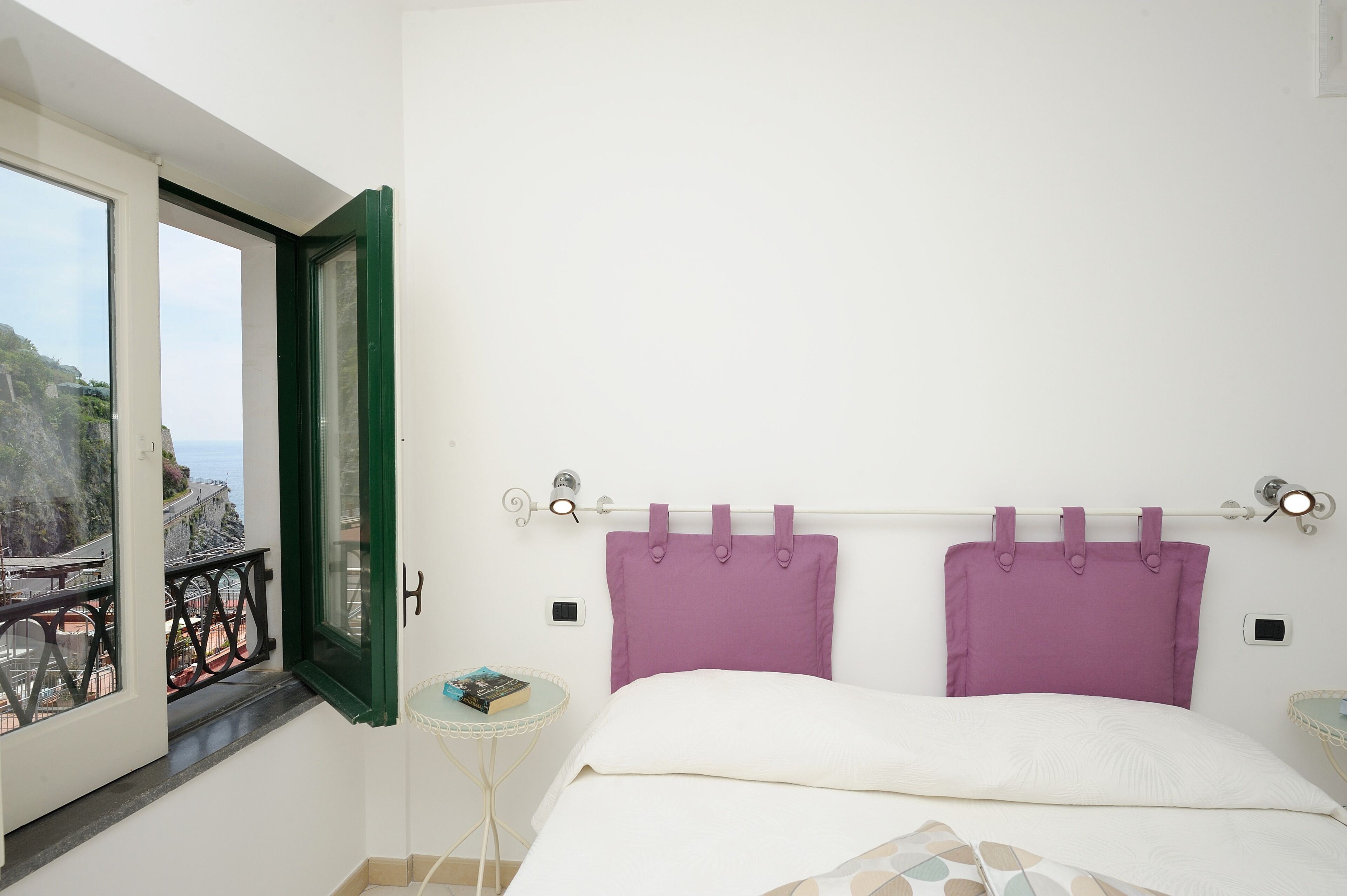 Un dormitorio con cama, ventana con vista al litoral, mesita de noche pequeña con un libro y otro mueble auxiliar.