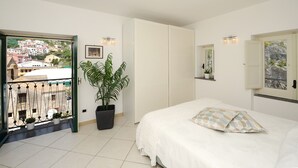 A bedroom with a bed, a potted plant, a window with a view of buildings, and a door. - Ananas - Sea View - Fruit Flats by AMALFIVACATION.IT (MINORI)