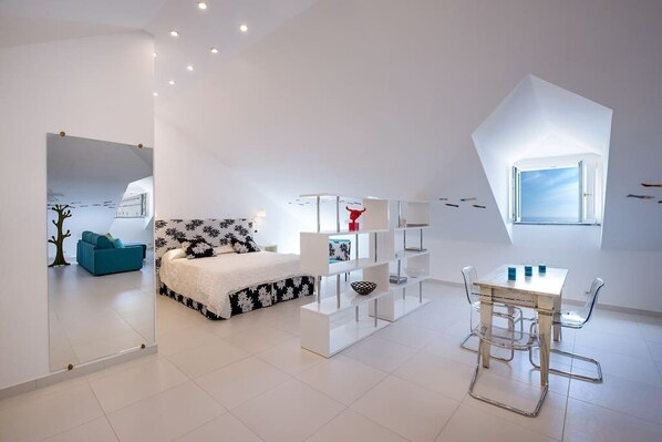 Une chambre à coucher moderne et minimaliste, avec un lit, une étagère blanche et un coin repas comprenant une table et des chaises.