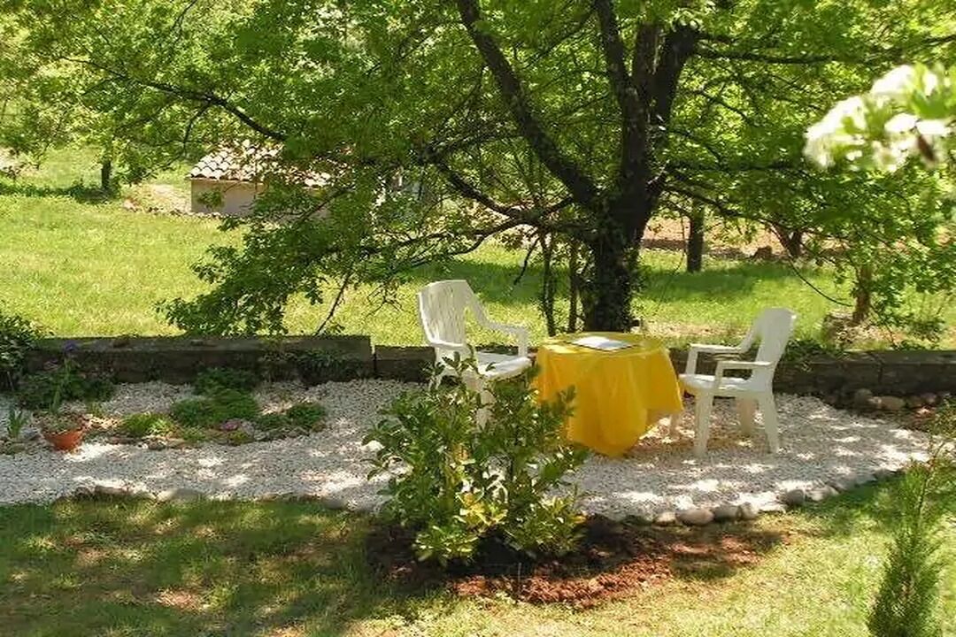 Ein kleiner Sitzbereich im Freien mit Tisch und Stühlen unter einem Baum, umgeben von einem Kiesweg und Grünflächen.