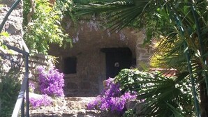 Eine Steintreppe führt zu einem rustikalen Gebäude mit einem kleinen Fenster, umgeben von üppigem Grün und violetten Blumen.