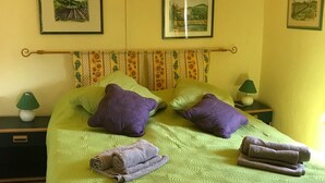 Ein ordentlich bezogenes Bett mit grüner Bettwäsche und violetten Kissen, ein Blumenmuster-Kopfschild sowie zwei gerahmte Bilder an der Wand.