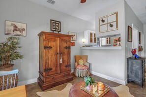 Una habitación con un armario grande de madera, una silla con un cojín con carita sonriente, una mesita con una vela y una planta en maceta.