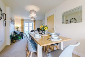 Un coin repas avec une table en bois dressée pour quatre personnes, entourée de chaises bleues. Un grand miroir orne le mur, et une horloge est placée au-dessus.