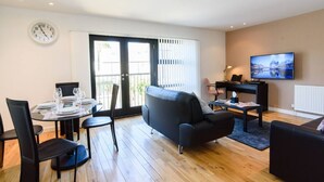 A modern living room with a black sofa, a dining table set for four, a TV, and a clock on the wall. - 15% OFF Superhost Stays Mn-Fri | Spacious 2 Bd Apt (Cardiff)