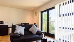 A modern living room with a black leather sofa, two patterned cushions, a magazine on the coffee table, and a window with blinds. - 15% OFF Superhost Stays Mn-Fri | Spacious 2 Bd Apt (Cardiff)