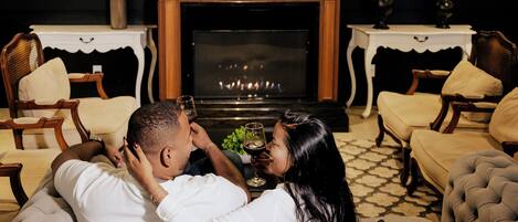 A couple relaxing on a tufted sofa in a room with a fireplace, wooden furniture, and a patterned floor.