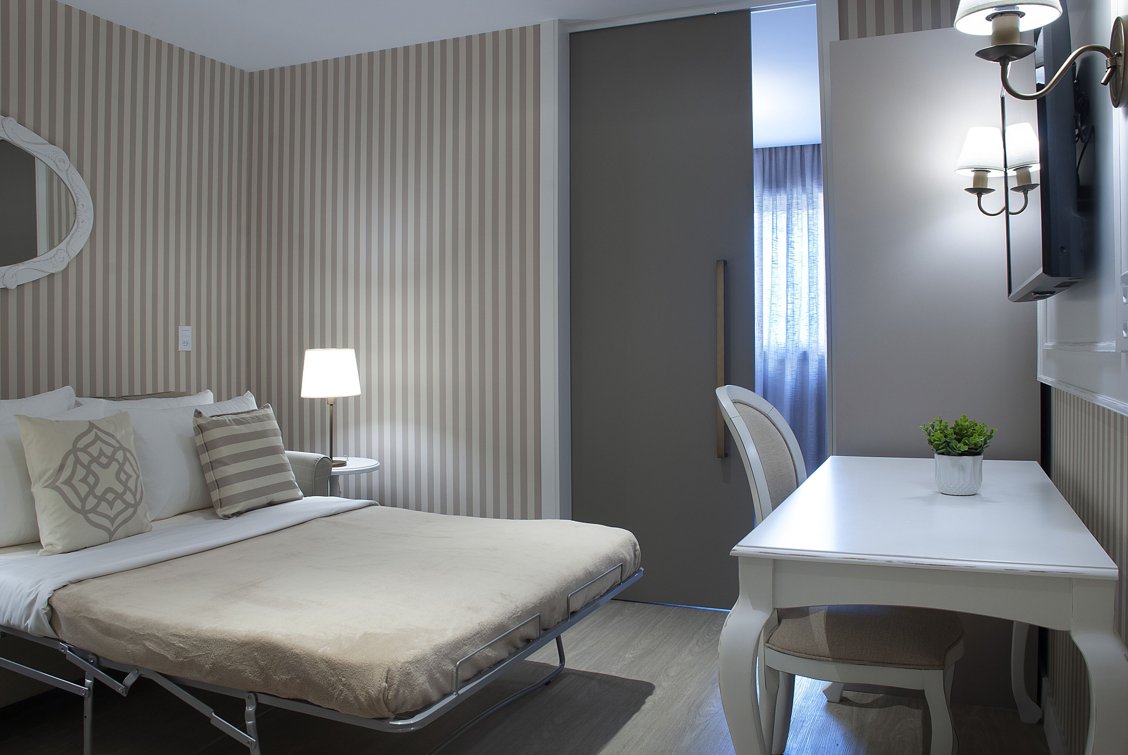 A hotel room with a bed, bedside table, lamp, chair, and a small table with a plant.