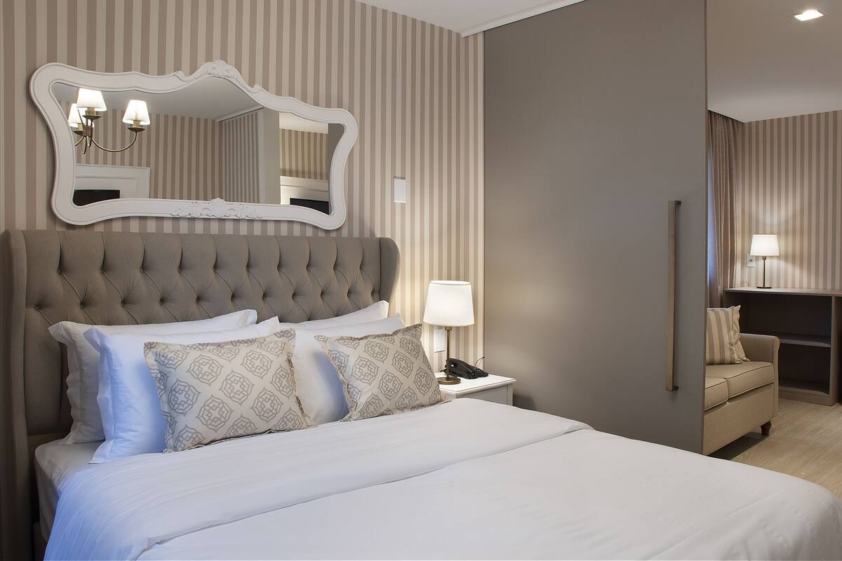 A neatly arranged hotel room with a large bed, a bedside table with a lamp, a chair, and a mirror on the wall.