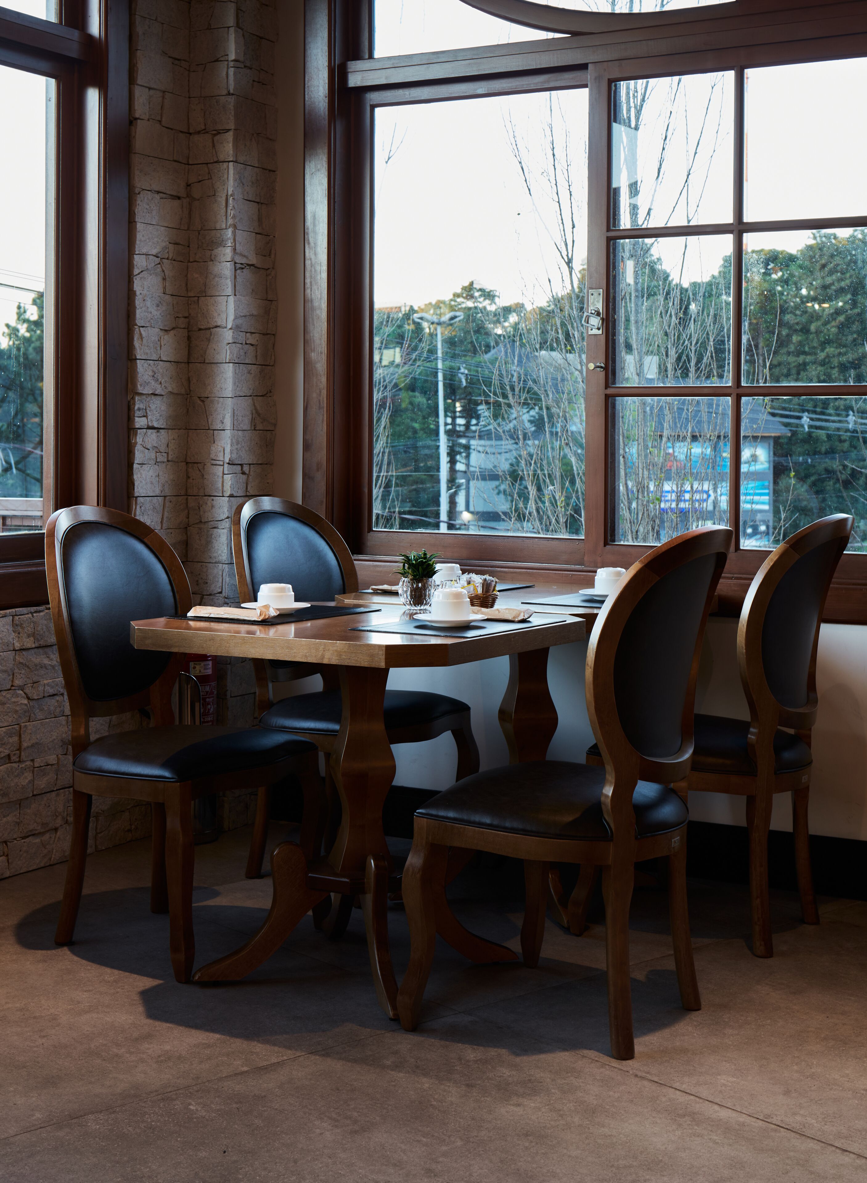 A dining area with a round wooden table surrounded by four chairs, a stone wall, and large windows with a view of trees and buildings.