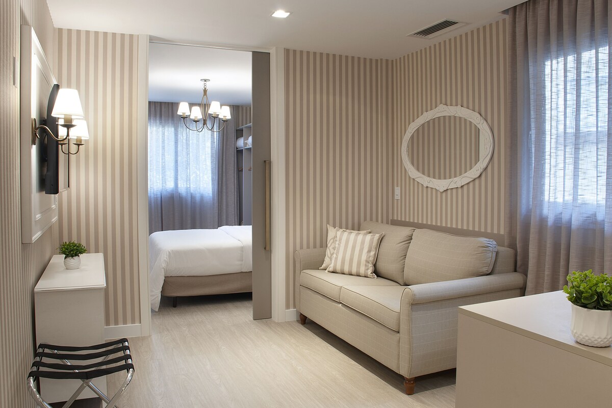 A hotel room with a bed, a sofa, a desk with a lamp, and a mirror on the wall.