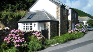 A white building with a stone facade and a sloped roof, surrounded by lush greenery and pink flowers. - 3 bedroom accommodation in Boscastle (Boscastle)