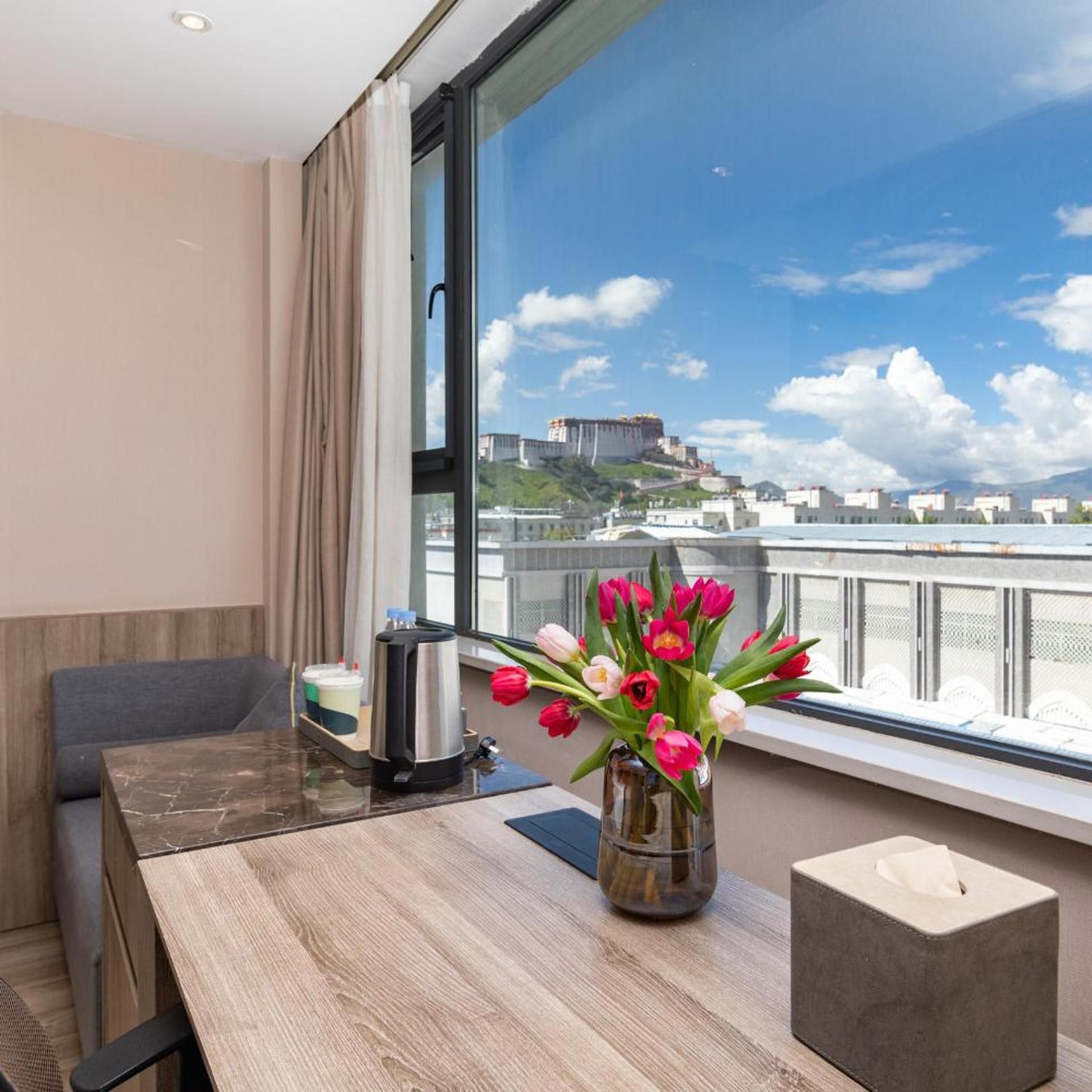 A hotel room with a large window, a table with a coffee maker, a vase of tulips, and a tissue box.