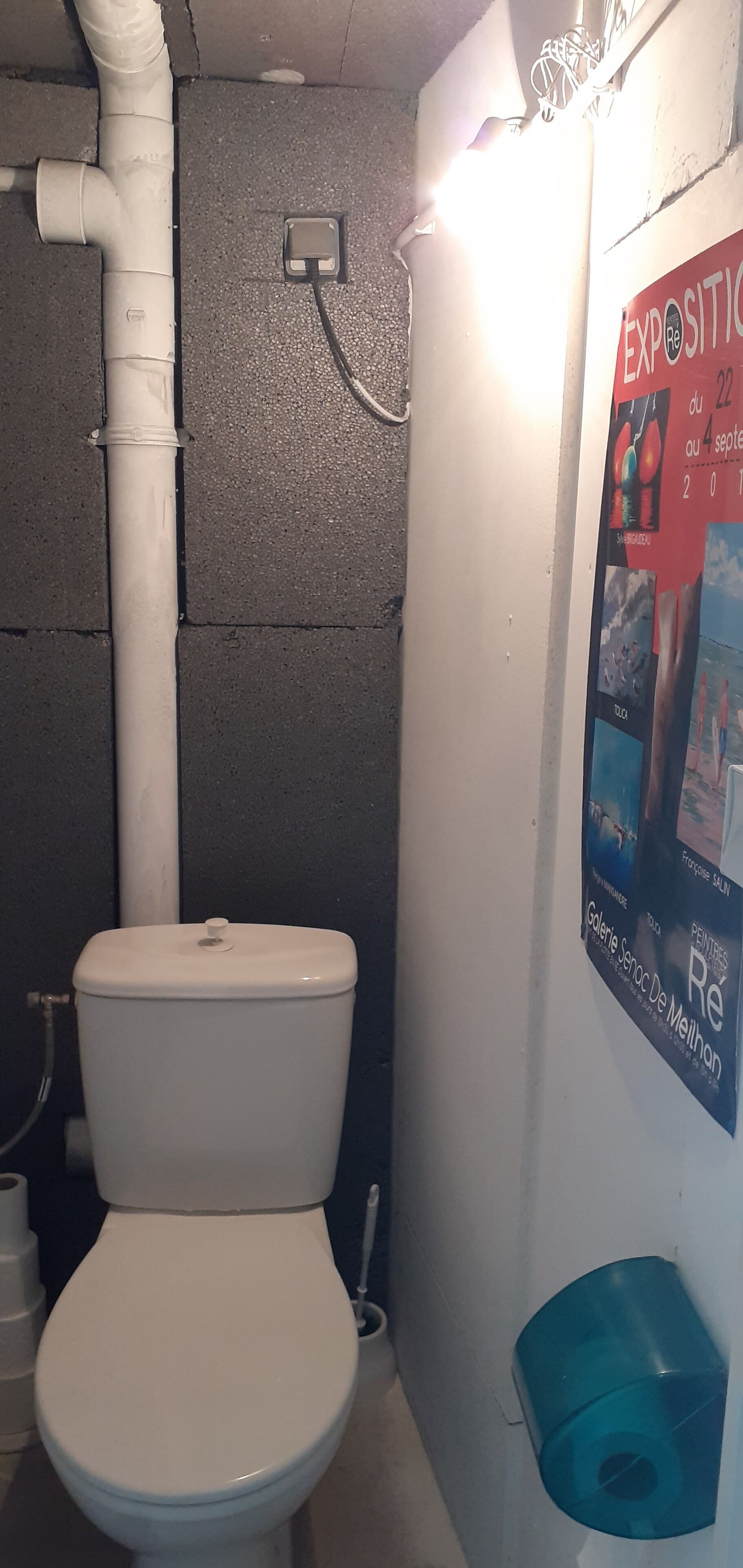 Un baño pequeño con un inodoro blanco, un bidé azul y un cartel en la pared que anuncia una exposición.