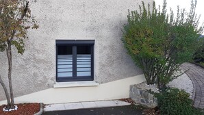 Un edificio con fachada texturizada, una ventana con persianas horizontales y un pequeño árbol en primer plano.