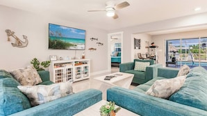 Een ruime woonkamer met blauwgroene banken, een salontafel en een televisie. Er is een eethoek met een tafel en stoelen, en er is uitzicht op een zwembad door grote ramen.