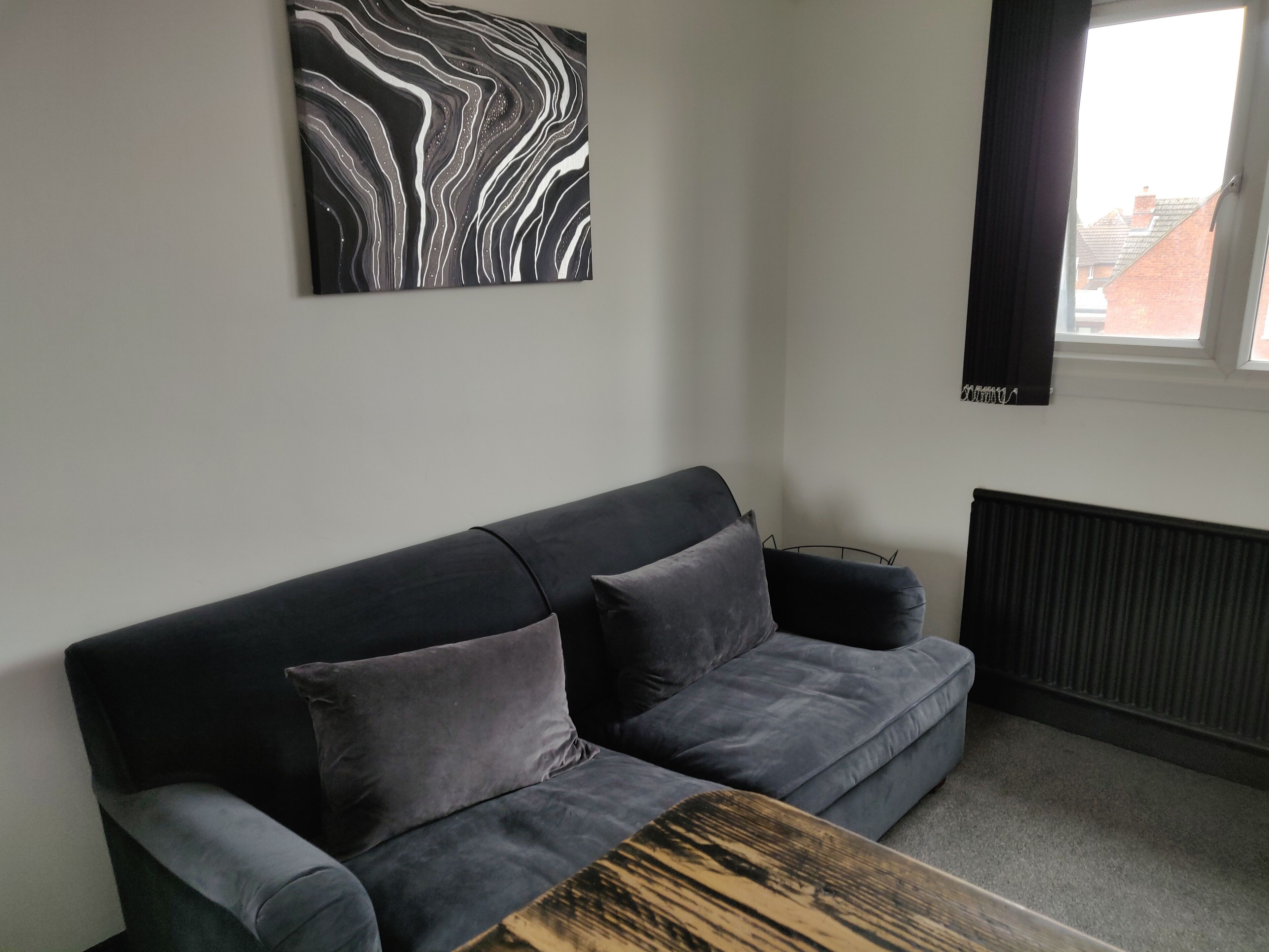 Een moderne woonkamer met een donkergrijze bank, twee kussens, een houten salontafel en een zwart-wit abstract schilderij aan de muur.