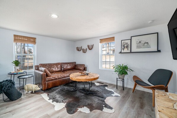 Una sala de estar moderna con un sofá de cuero, una mesa redonda de centro y un tapiz de cuero de vaca. Hay dos ventanas con persianas, un estante montado en la pared con obras de arte y una mesita auxiliar con una planta.