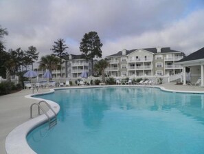 A swimming pool with lounge chairs and umbrellas, surrounded by multi-story buildings with balconies. - Calabash, NC - 1BD/1BA Condo 1st Floor Unit - Near NC/SC Beaches & Golf Courses (Calabash)