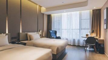 A hotel room with two beds, a desk, a chair, and a large window with curtains.