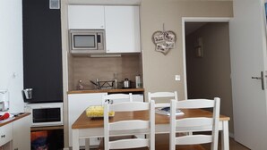 Een kleine keuken met witte kastjes, een magnetron en een spoelbak. Er staat een eettafel met witte stoelen en een kom met ontbijtgranen.