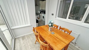 Een eetgedeelte met een houten tafel en stoelen, een kleine vaas met bloemen, en uitzicht op de keuken door een geopende deur.