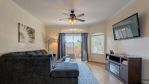 A living room with a sectional sofa, a flat-screen TV, a ceiling fan, and a large window with curtains. - Resort Style Living in Prime Scottsdale Location (Scottsdale)